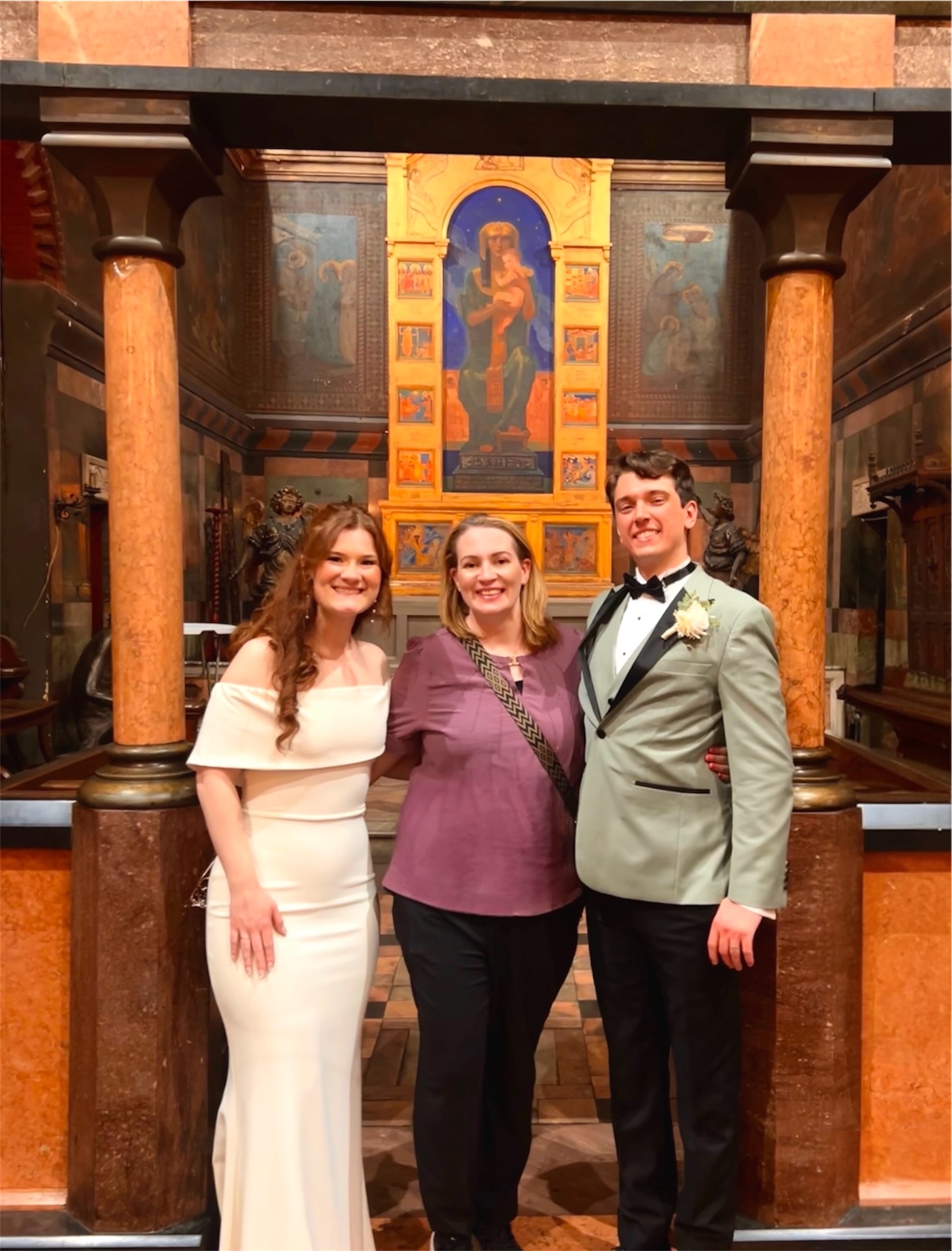 Three people are standing together in an ornate room with religious artwork on the wall behind them. The person on the left is a woman in a white dress, the person in the middle is a woman in a purple top, and the person on the right is a man in a gray tuxedo with a black bow tie.