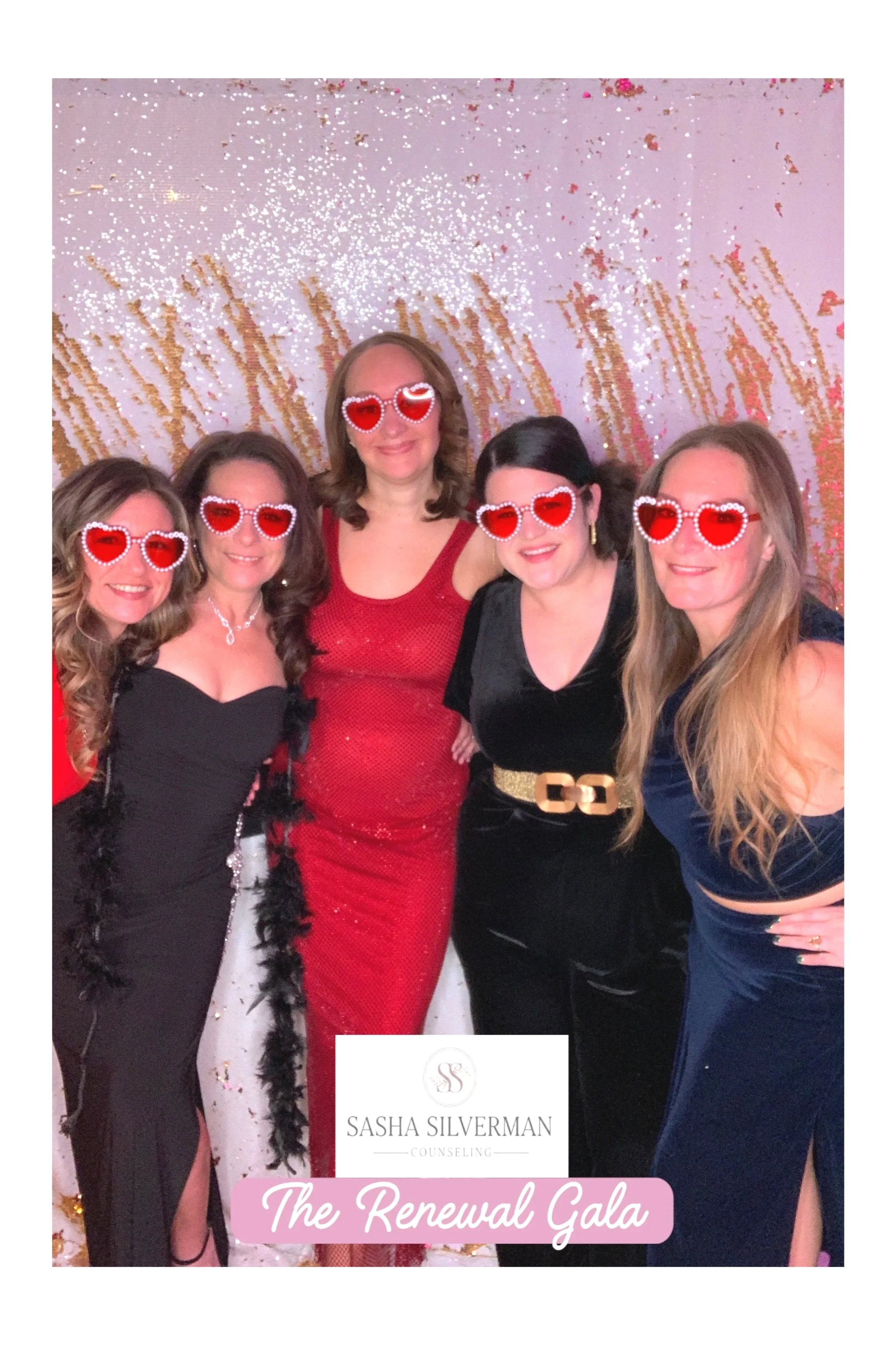 Group of five women wearing heart-shaped sunglasses with red lenses, dressed in elegant evening attire, at an event called The Renewal Gala sponsored by Sasha Silverman Counseling, standing in front of a gold and pink sparkling backdrop.