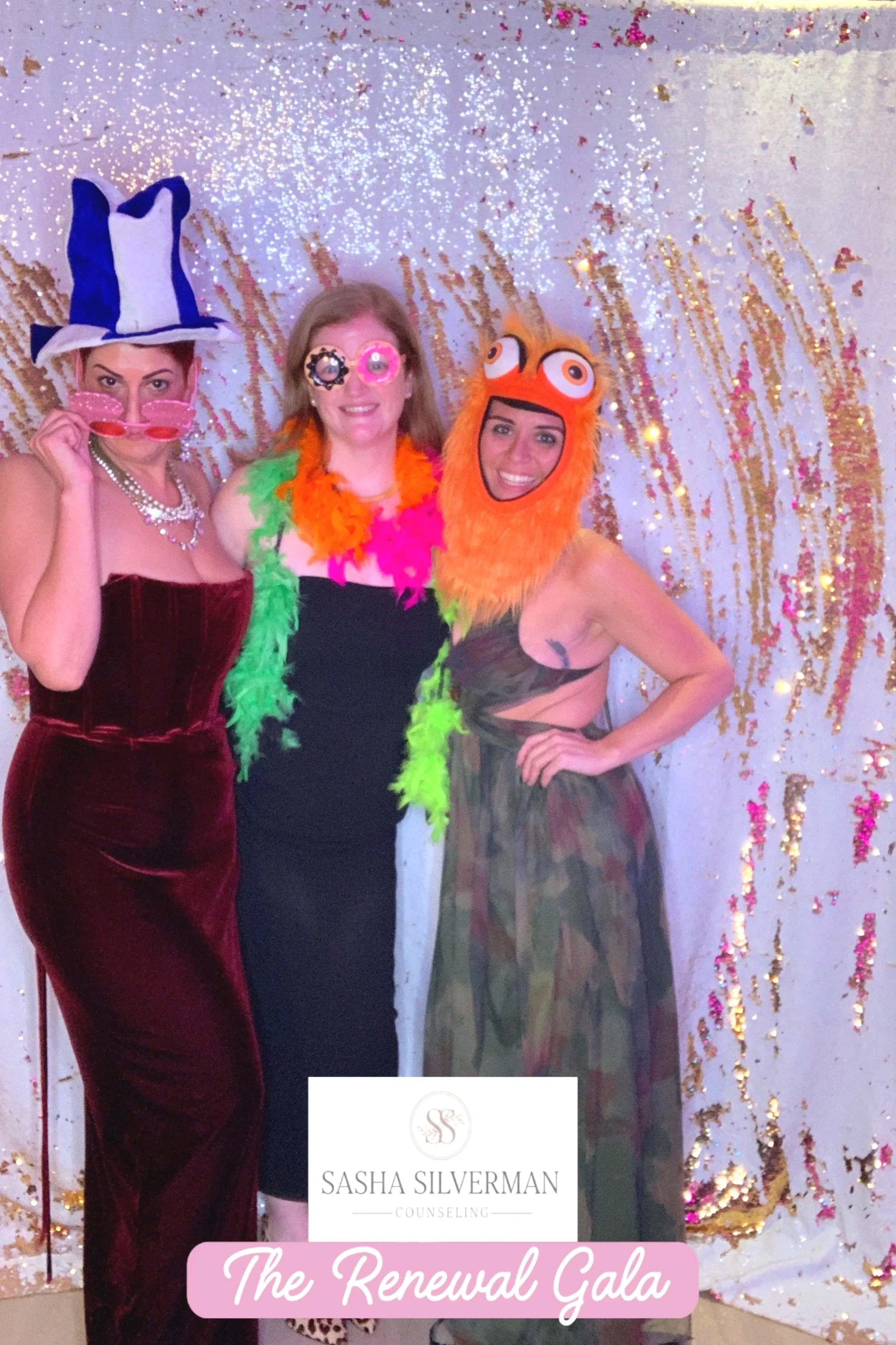 Three women at a festive event wearing colorful costumes and props, standing in front of a glittery backdrop with gold and pink decorations, promoting Sasha Silverman Counseling at 'The Renewal Gala'.