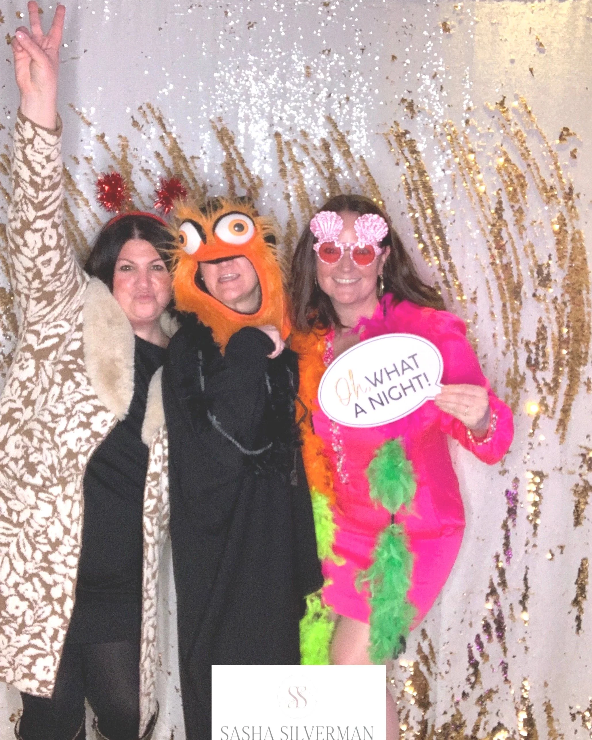 Three women dressed in colorful costumes celebrating at a party. One woman is wearing a leopard print coat with red headband antlers, another is wearing a monster costume with an orange furry face, and the third is in a bright pink dress with shell g