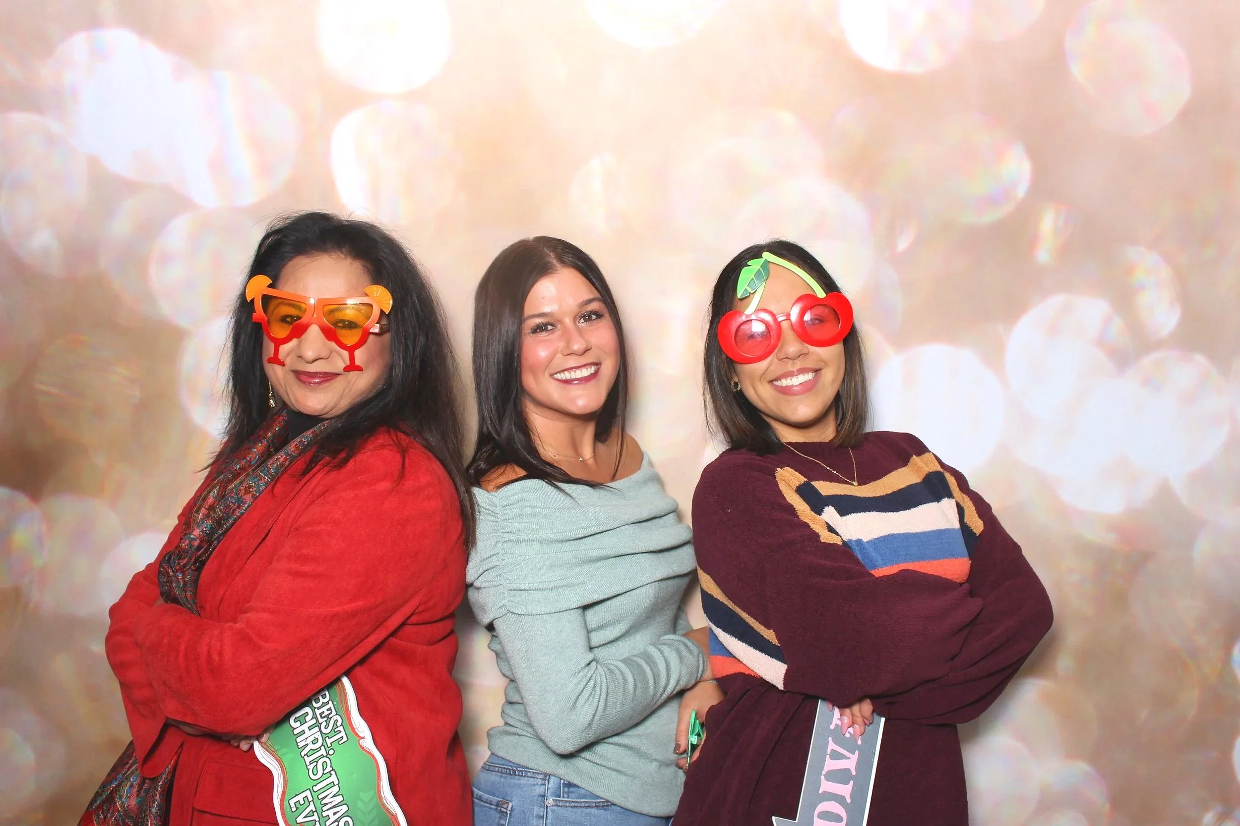 Three women posing together at a festive event, wearing colorful glasses and smiling. They are standing against a background with bokeh lights.