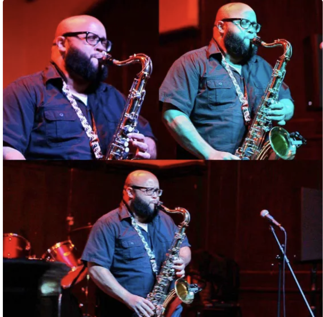 A man with glasses and a beard playing a saxophone on stage in a dimly lit venue, in front of a microphone stand.