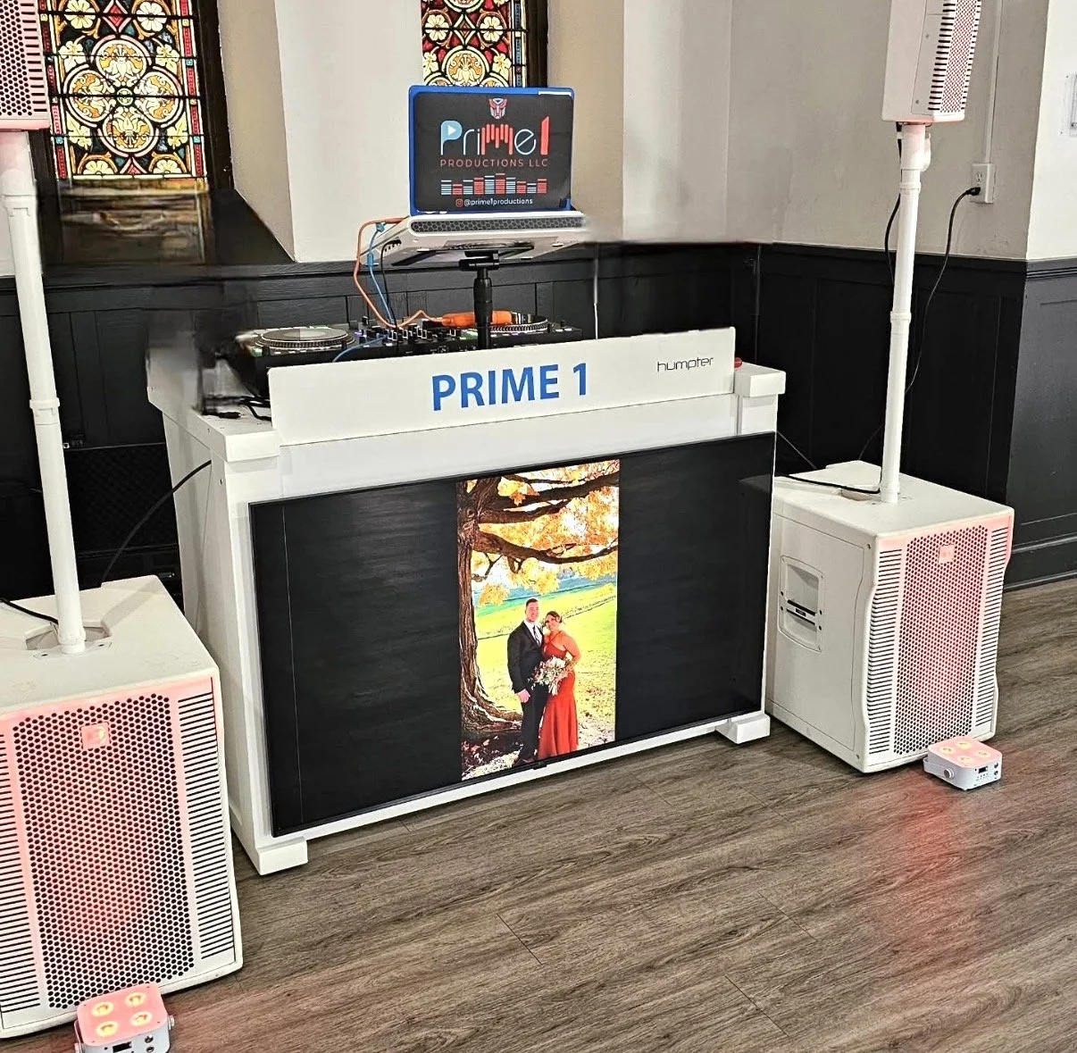 A DJ setup with speakers, a laptop on a stand, and a large screen displaying a wedding photo of a couple standing under a tree with autumn leaves. The DJ booth has "PRIME 1" written on it, and there are two pink-colored marshall speakers on either si