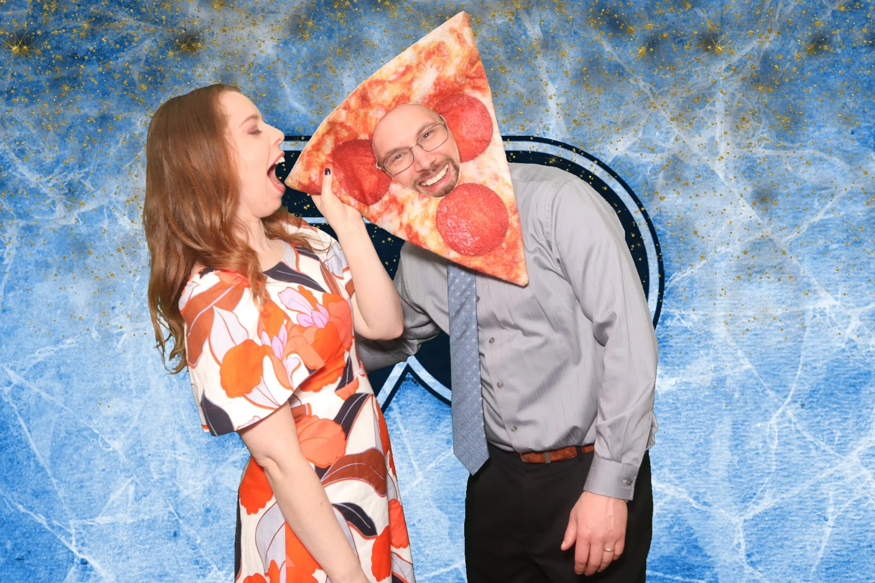 A woman is playfully pretending to bite a man wearing a pizza slice costume with pepperoni. The background is a textured blue with a gold splatter pattern and a shield emblem.