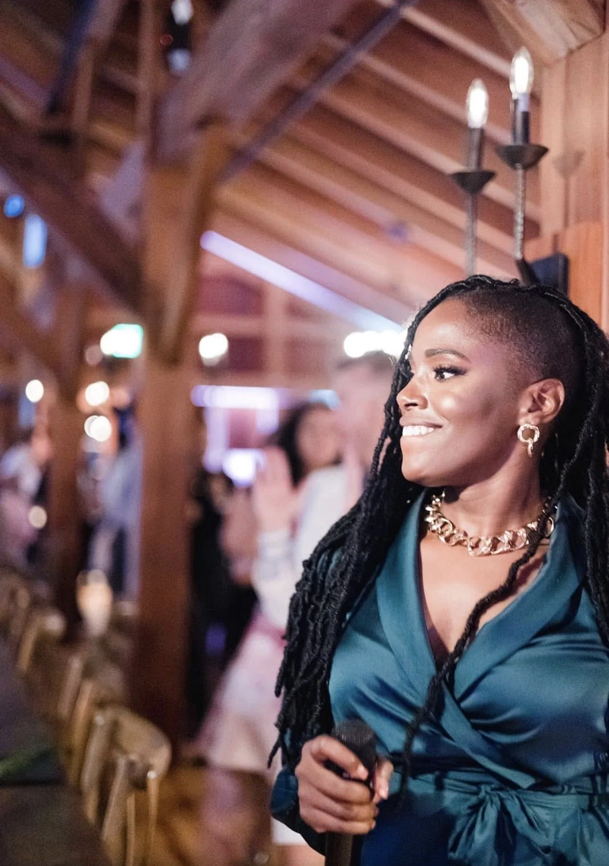 A woman with long, styled dreadlocks smiling at an indoor event. She is wearing a teal top, gold jewelry, and holding a microphone, with blurred background of people and wooden decor.