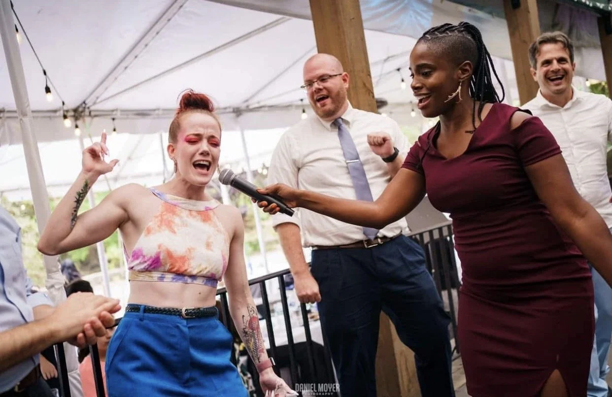 A group of people at a celebration under a tent, with a woman singing into a microphone, others smiling and enjoying the moment.