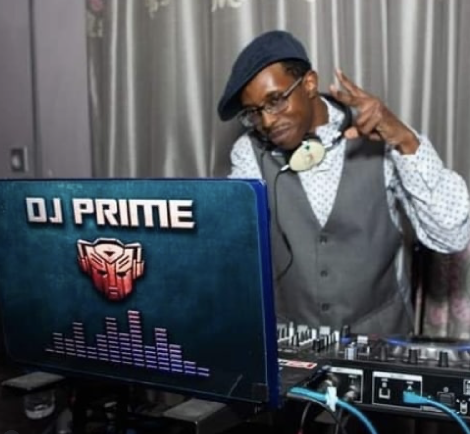 A man wearing a black beret, glasses, and a grey vest with a white shirt making a peace sign with his right hand while DJing at a setup with a monitor that displays DJ PRIME and a Transformers logo.