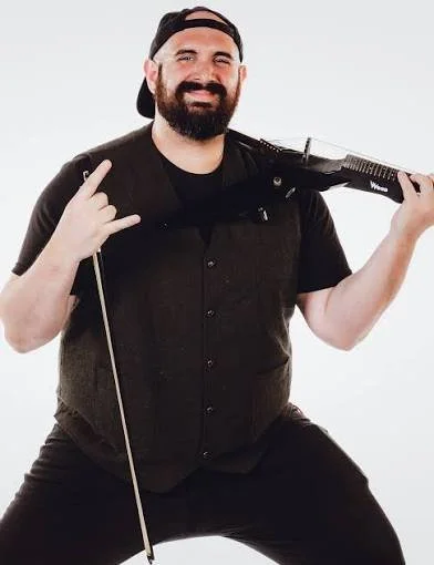 A man with a beard wearing a baseball cap backwards, a black t-shirt, and a vest, smiling and posing with a weapon over his shoulder. He is making a rock and roll hand gesture with his left hand.