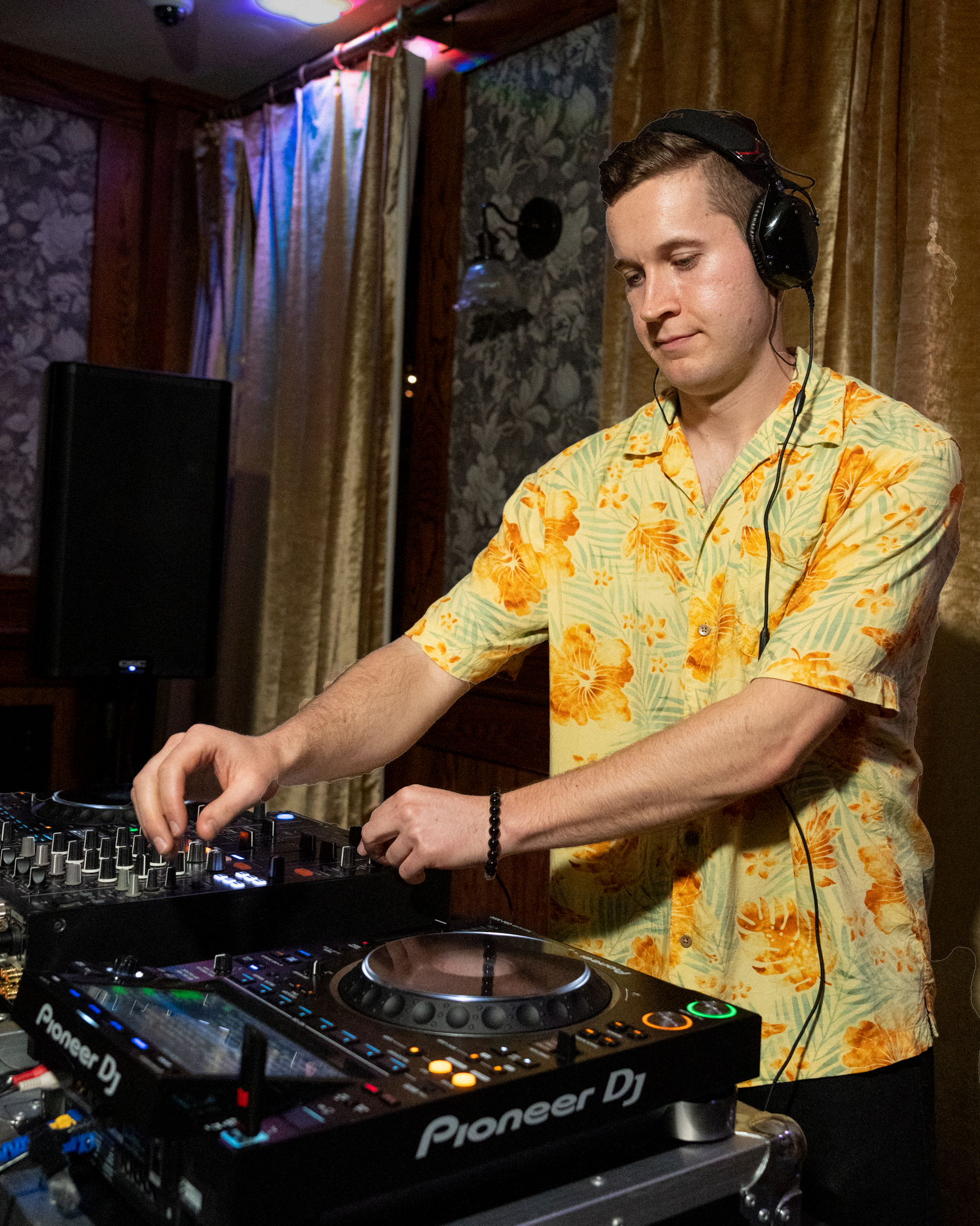DJ Zach Atom wearing a yellow Hawaiian shirt with orange floral patterns, with headphones on, is operating a DJ mixer in a dimly lit room with floral wallpaper and gold curtains.