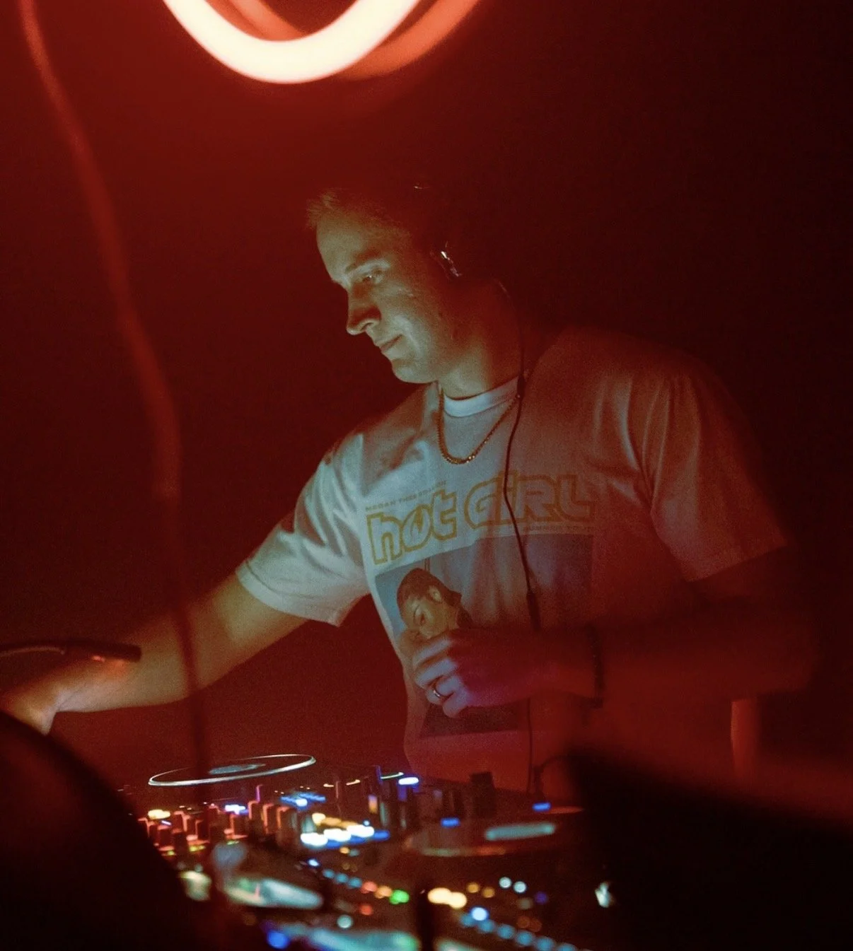 A DJ wearing a Megadeth t-shirt, headphones, and a necklace, working at a DJ console in a dimly lit environment with red lighting.