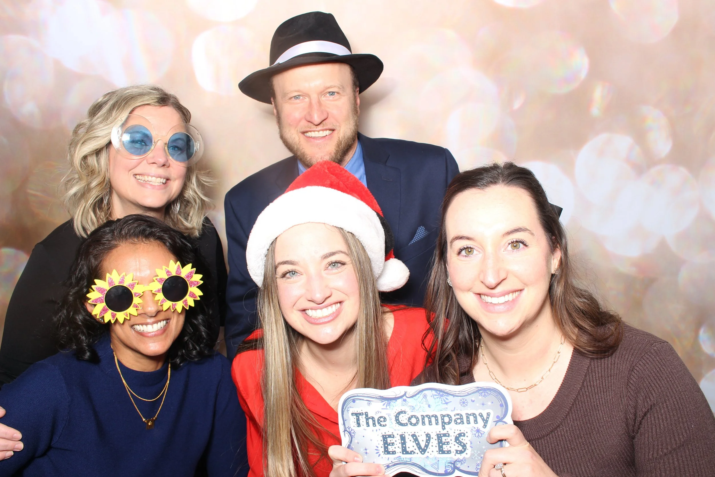A group of six people celebrating Christmas and New Year, wearing festive accessories such as a Santa hat, Christmas hat, funky glasses, and a fedora. They are smiling and holding a sign that reads 'The Company Elves,' with a festive background of bl
