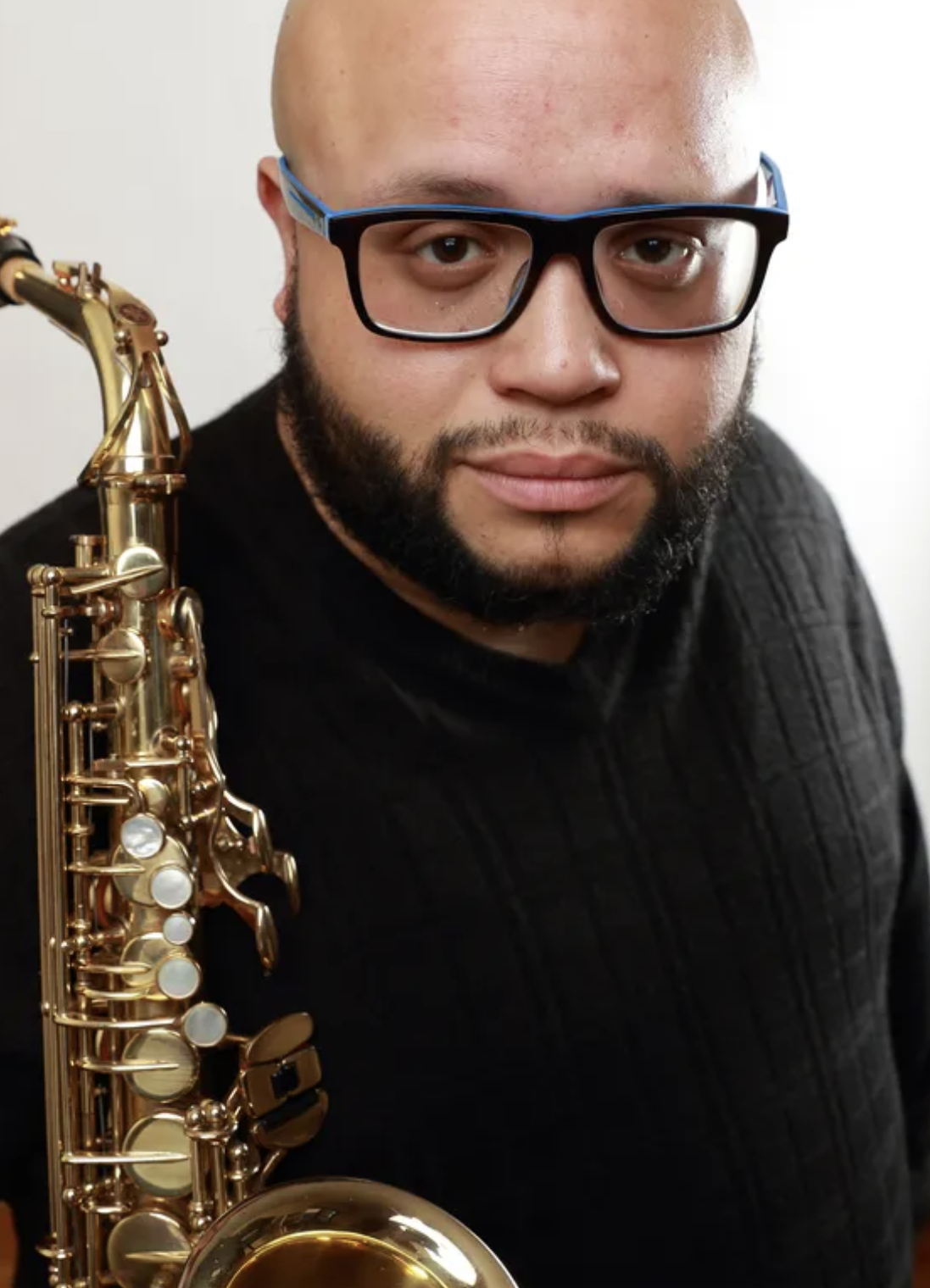 A man with glasses and a beard holding a gold saxophone, wearing a black sweater, against a plain white background.