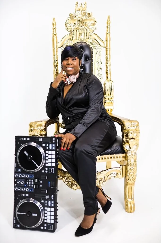 A woman with short black hair, dressed in black, sitting on a gold and black throne-like chair, wearing headphones, with a DJ controller on the floor next to her, smiling at the camera.