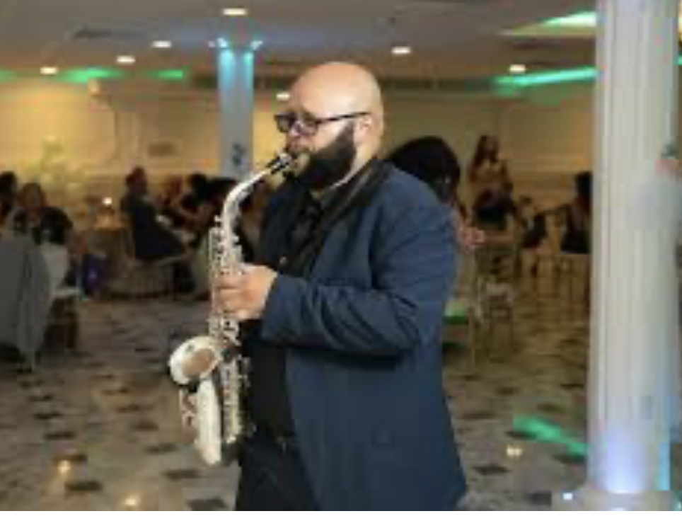A man with a bald head, glasses, and a beard playing a saxophone in a dimly lit indoor venue with green and blue decorative lighting, and people sitting at tables in the background.