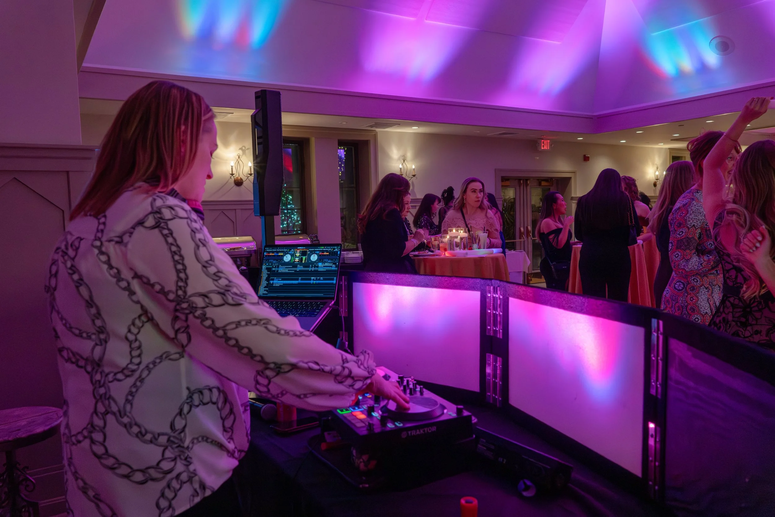 A woman DJ is performing at an event with women dancing and socializing in the background. The room is decorated with colorful lighting and a festive atmosphere.