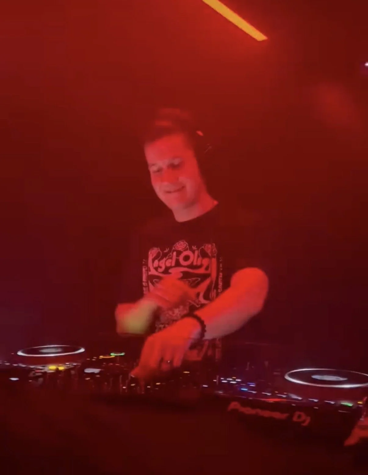A male DJ with short dark hair, wearing a black T-shirt with pink and white graphics, is performing at a club with red lighting. He is smiling while operating DJ equipment on a table.