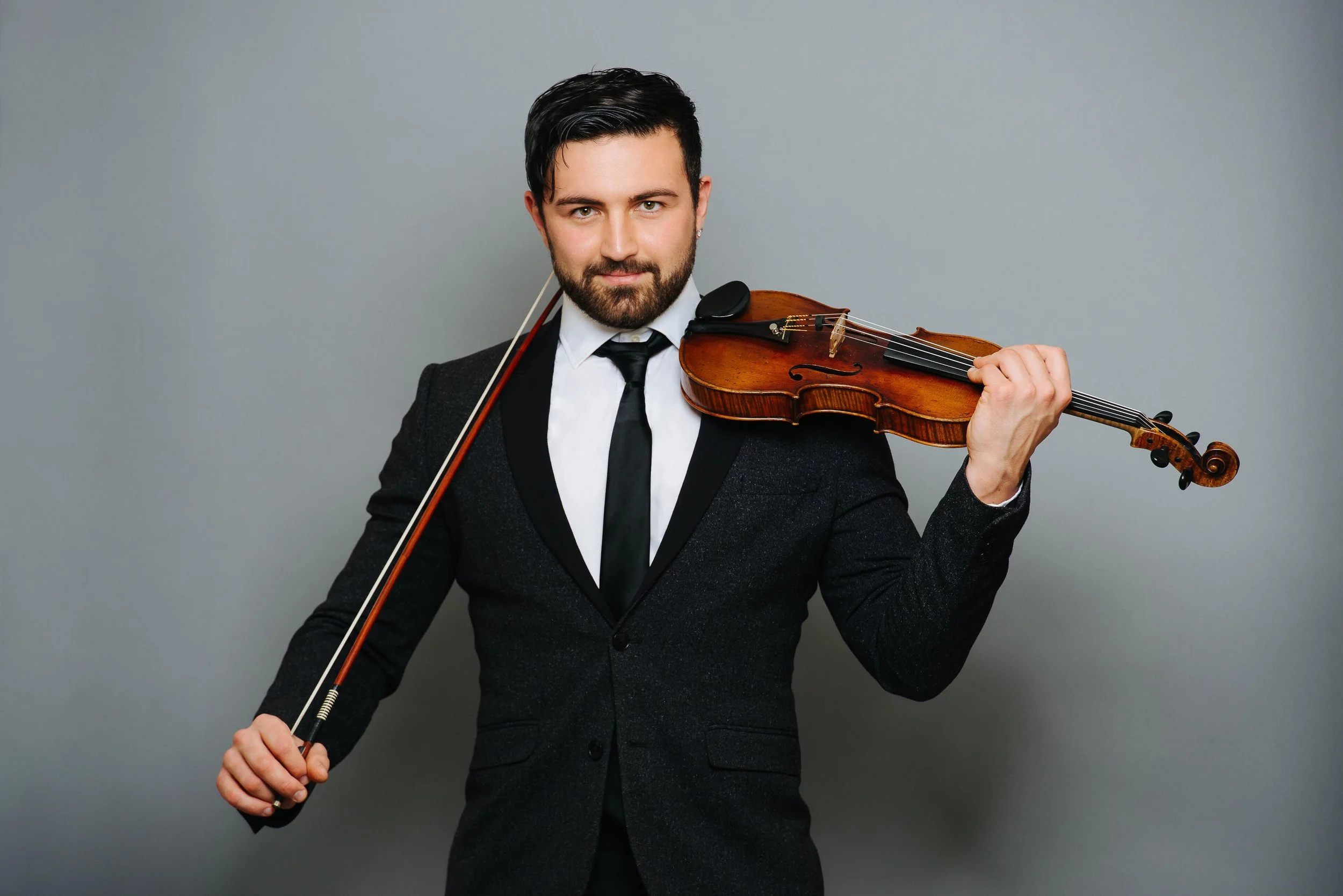 A man in a dark suit and tie holding a violin and bow against a gray background.