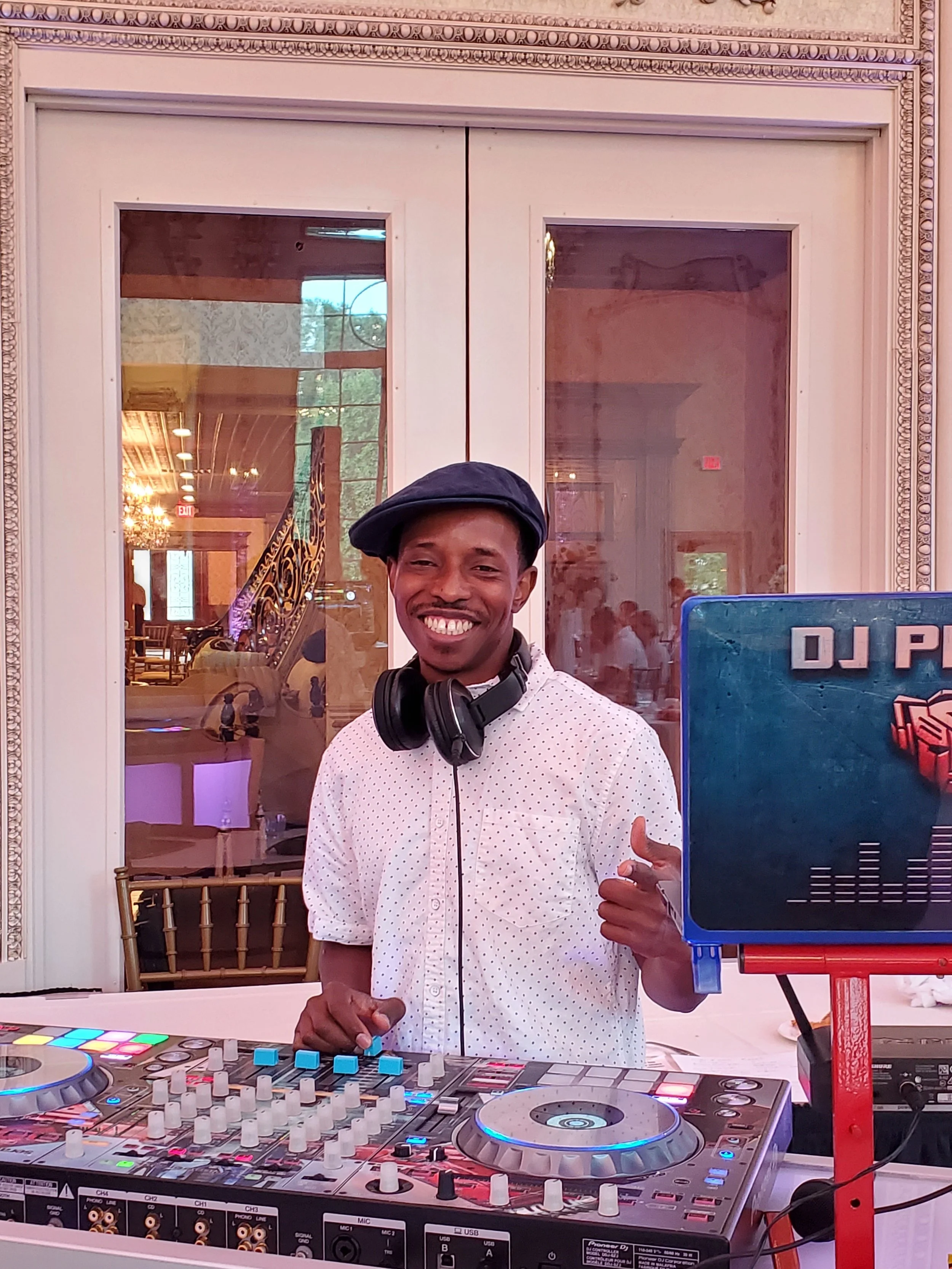 A smiling DJ standing behind a DJ console at an indoor event, wearing a white polka dot shirt, black cap, and headphones around his neck, with a blue DJ sign beside him.