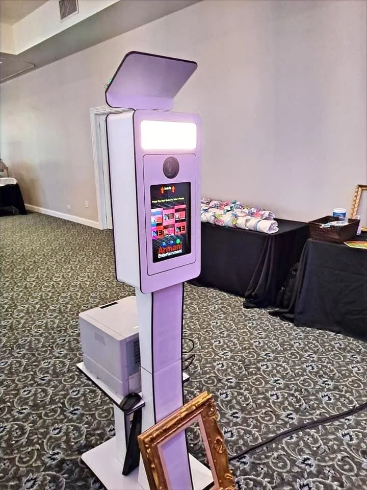 A purple photo booth with a digital screen, top open cover, and a framed empty picture leaning against its base, set up indoors on a patterned carpet.