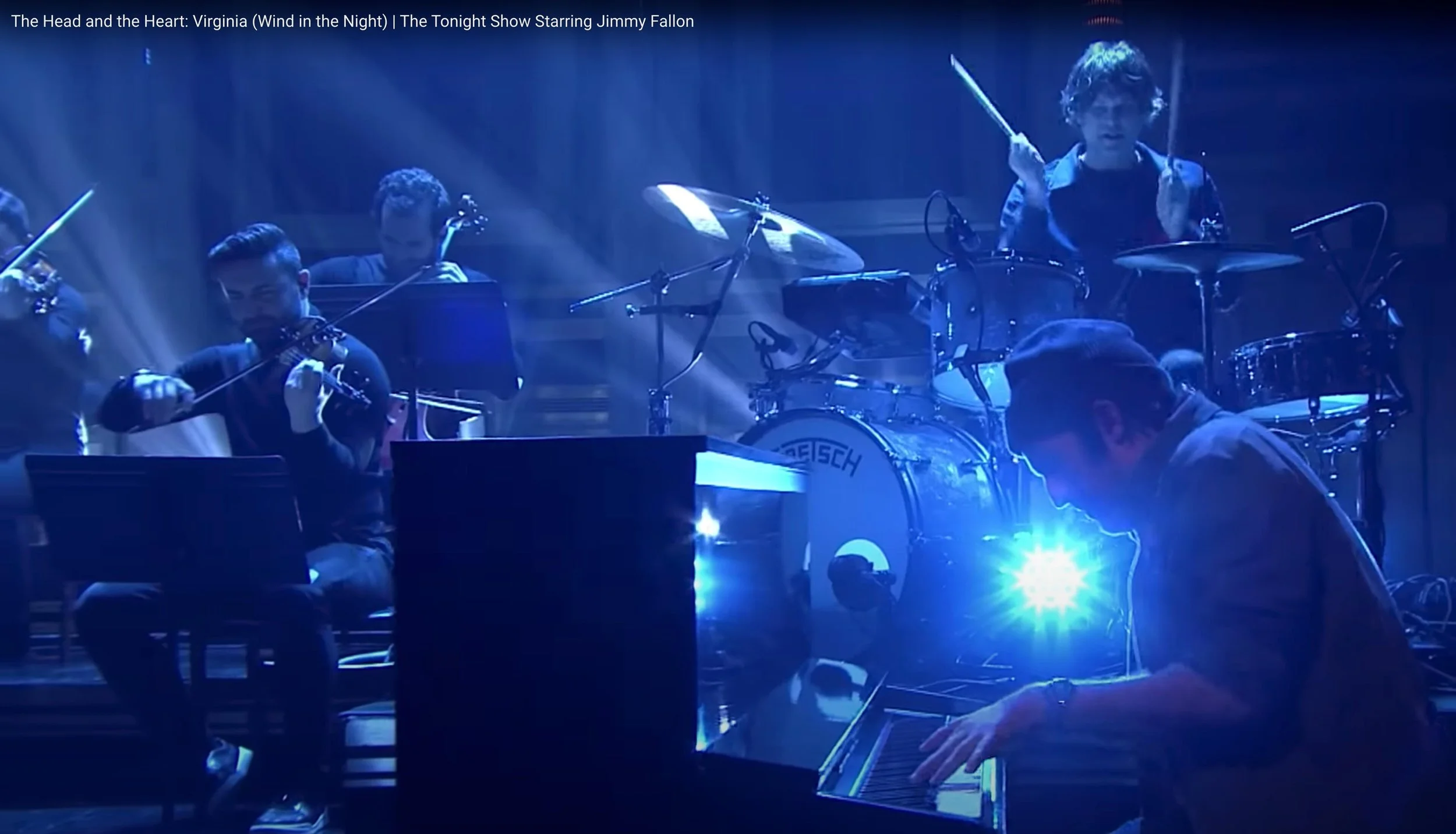 A group of musicians performing on stage with blue lighting, including a young drummer and a violinist, and a pianist in the foreground.