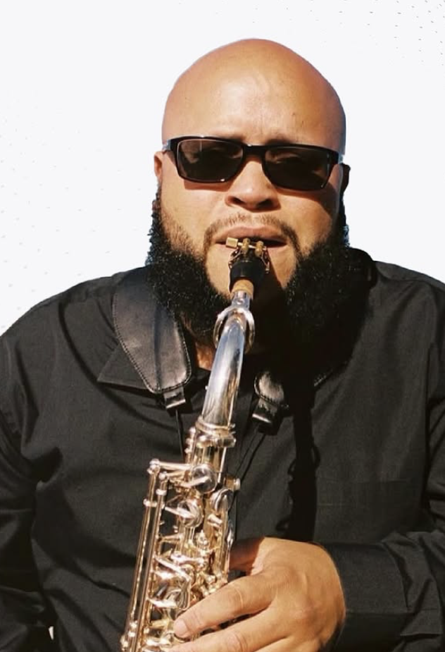A man with a beard wearing black sunglasses and a black jacket, playing a saxophone.