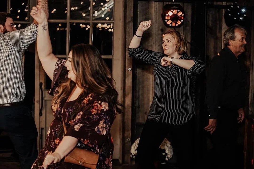 People dancing and enjoying themselves at a social gathering or party, with fairy lights and rustic decor in the background.