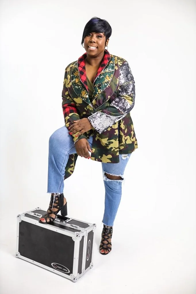 A confident woman wearing a camo jacket, ripped jeans, and black high-heeled sandals, standing with one foot on a black equipment case against a plain white background.