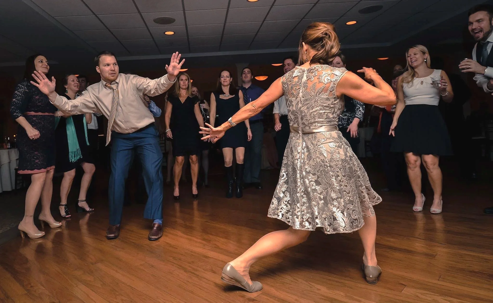 The bride and groom dancing in the center of a group of people at a party or celebration, with some onlookers clapping and smiling on a the dance floor.