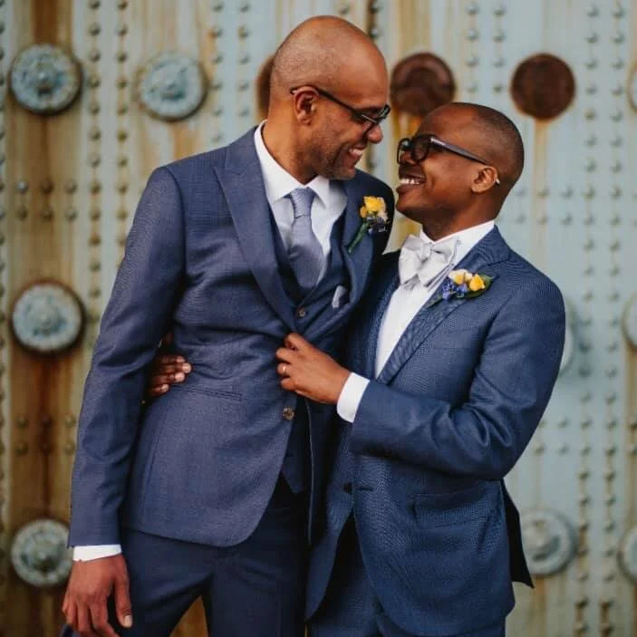 Two grooms smiling and looking at each other, standing close with one man's arm around the other's waist. They are having a photo shoot at their Philadelphia wedding, with a decorated backdrop of hanging plates and wallpaper.