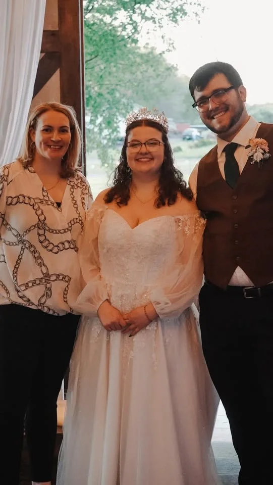 Three people at a wedding, including a bride in a white wedding gown with a tiara, a woman in a patterned blouse, and a man in a vest, tie, and boutonniere, standing inside near a window with greenery outside.