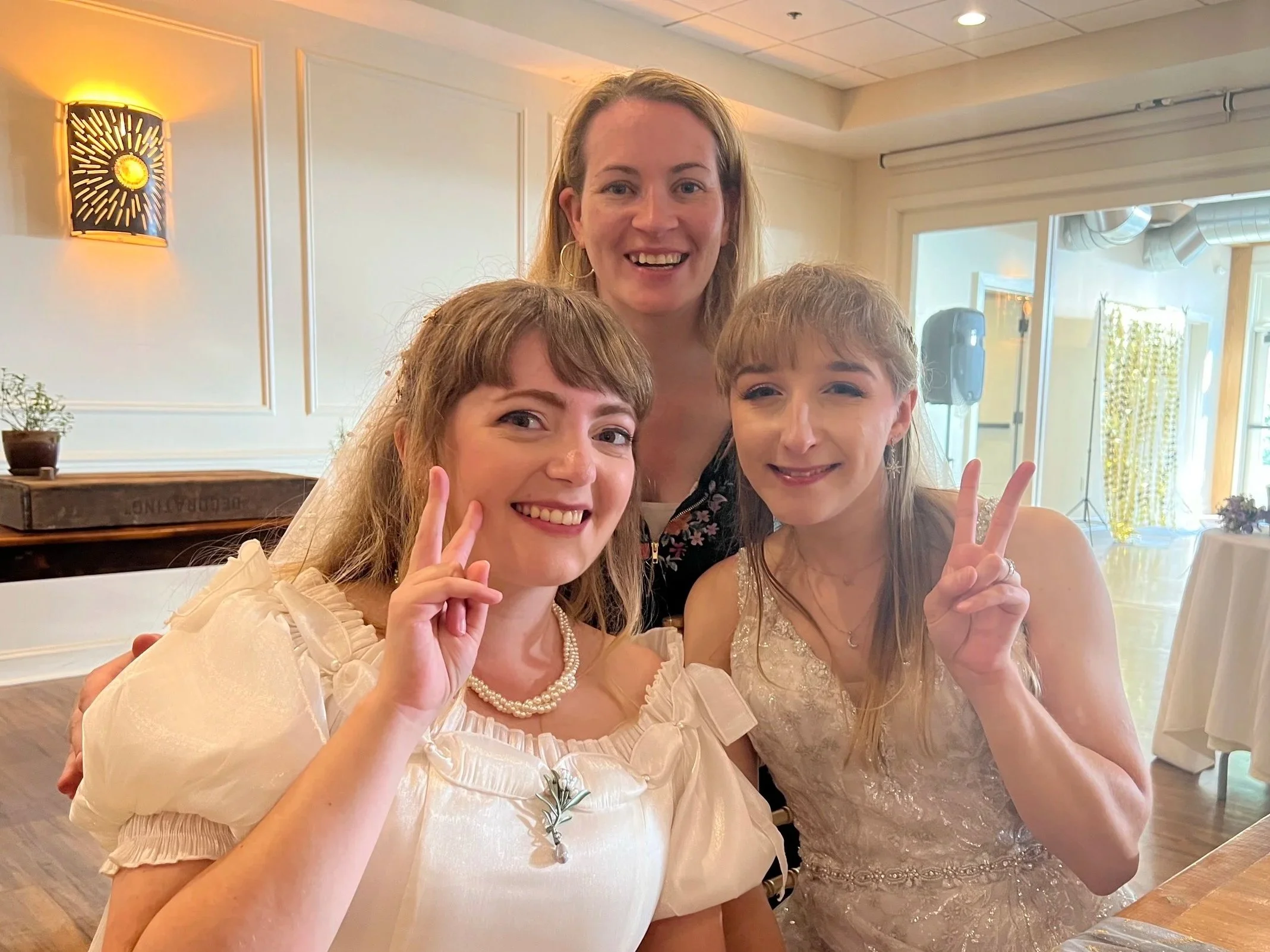 Three women, two in wedding dresses and one in a black floral top, smiling and making peace signs in a brightly lit room during a celebration.
