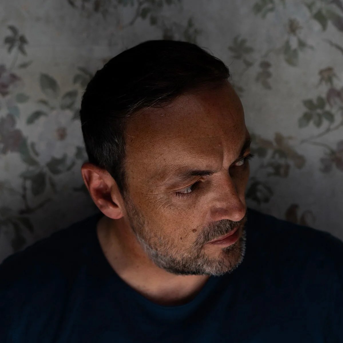 Close-up d'un homme avec une barbe, regardant vers le bas, avec un fond de papier peint à motifs floraux.
