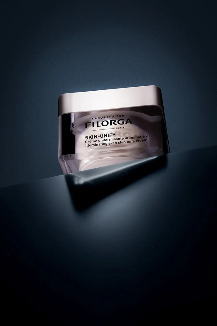 Pot de crème pour la peau Filorga Skin-Unify, placé sur une surface sombre, avec un fond dégradé