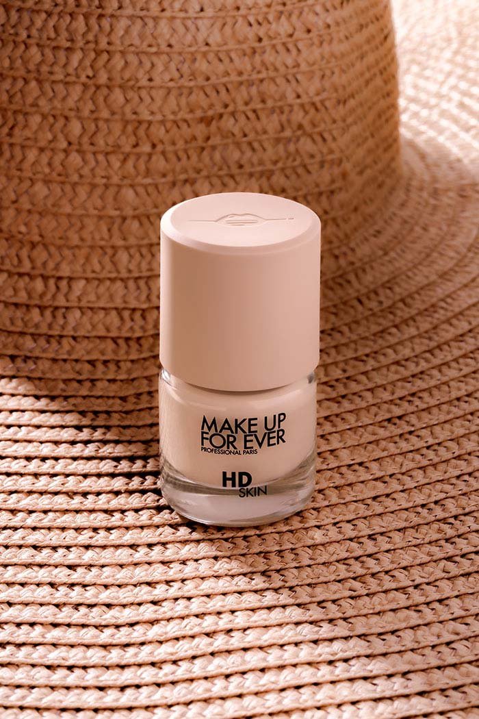 Vernis à ongles blanc de la marque Make Up For Ever, posé sur un tapis en rotin avec un chapeau en paille en arrière-plan.