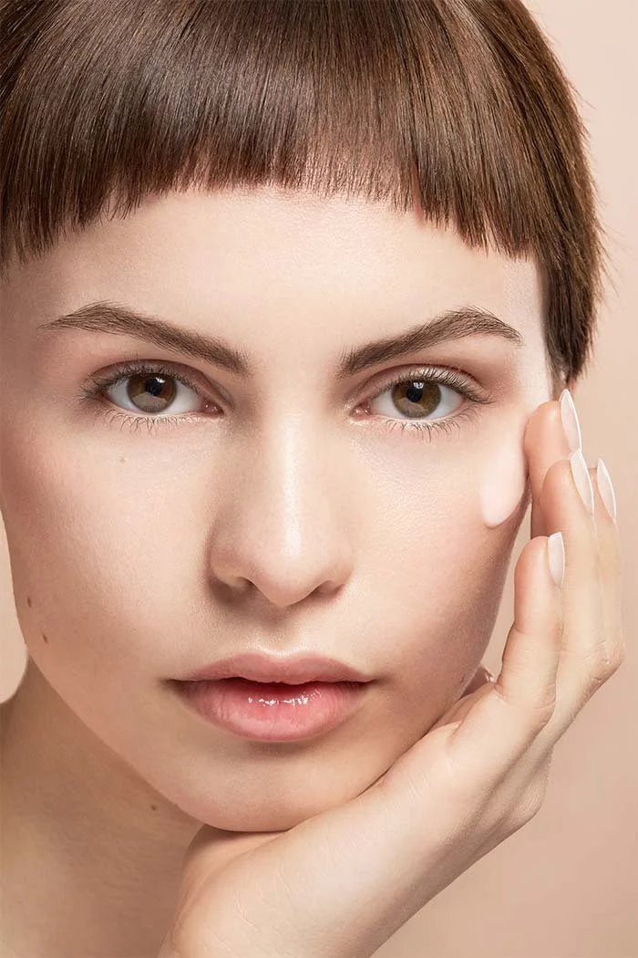 Portrait d'une femme avec une peau claire et des cheveux courts bruns, touchant doucement sa joue avec ses doigts.