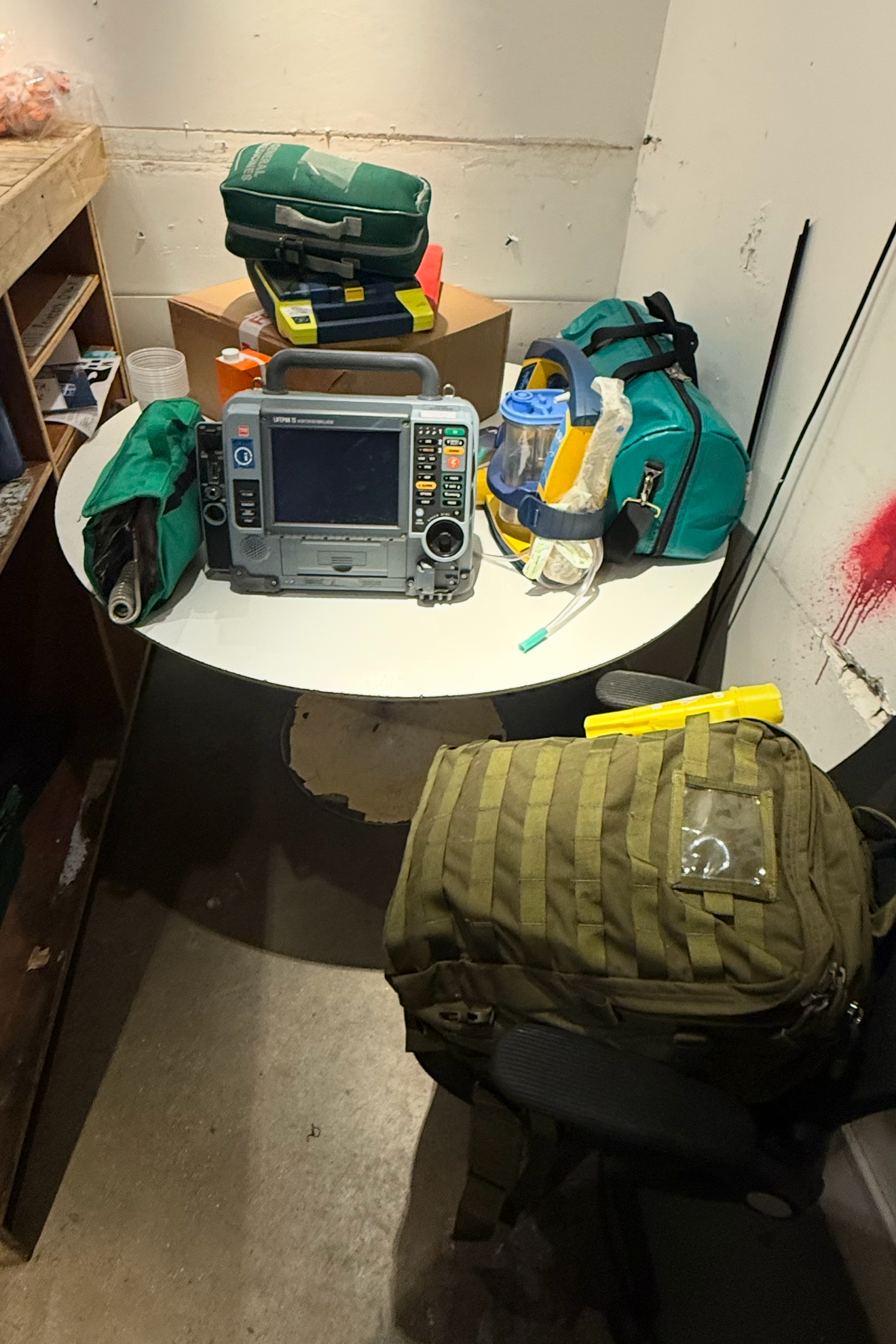 Emergency medical equipment and supplies on a table and chair, including a defibrillator, medical bags, protective visor, and tools.