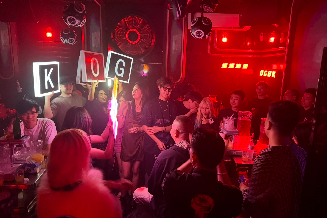 People dancing and socializing in a dimly lit nightclub with red lighting, holding illuminated signs spelling 'K', 'O', and 'G', surrounded by drinks and music equipment.