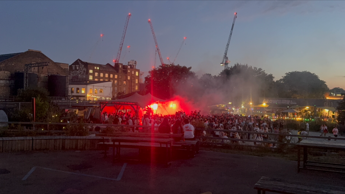 A crowd gathered outdoors at dusk with a stage illuminated in red light, smoke, and three construction cranes in the background.