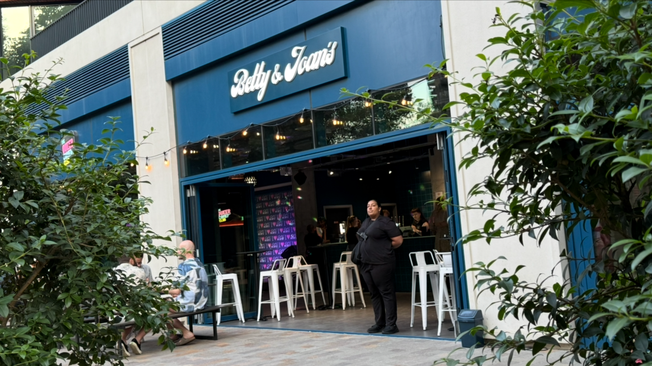 People sitting outside the Betty & Joan's restaurant, with some engaged in conversation and others waiting, while a woman stands near the entrance.