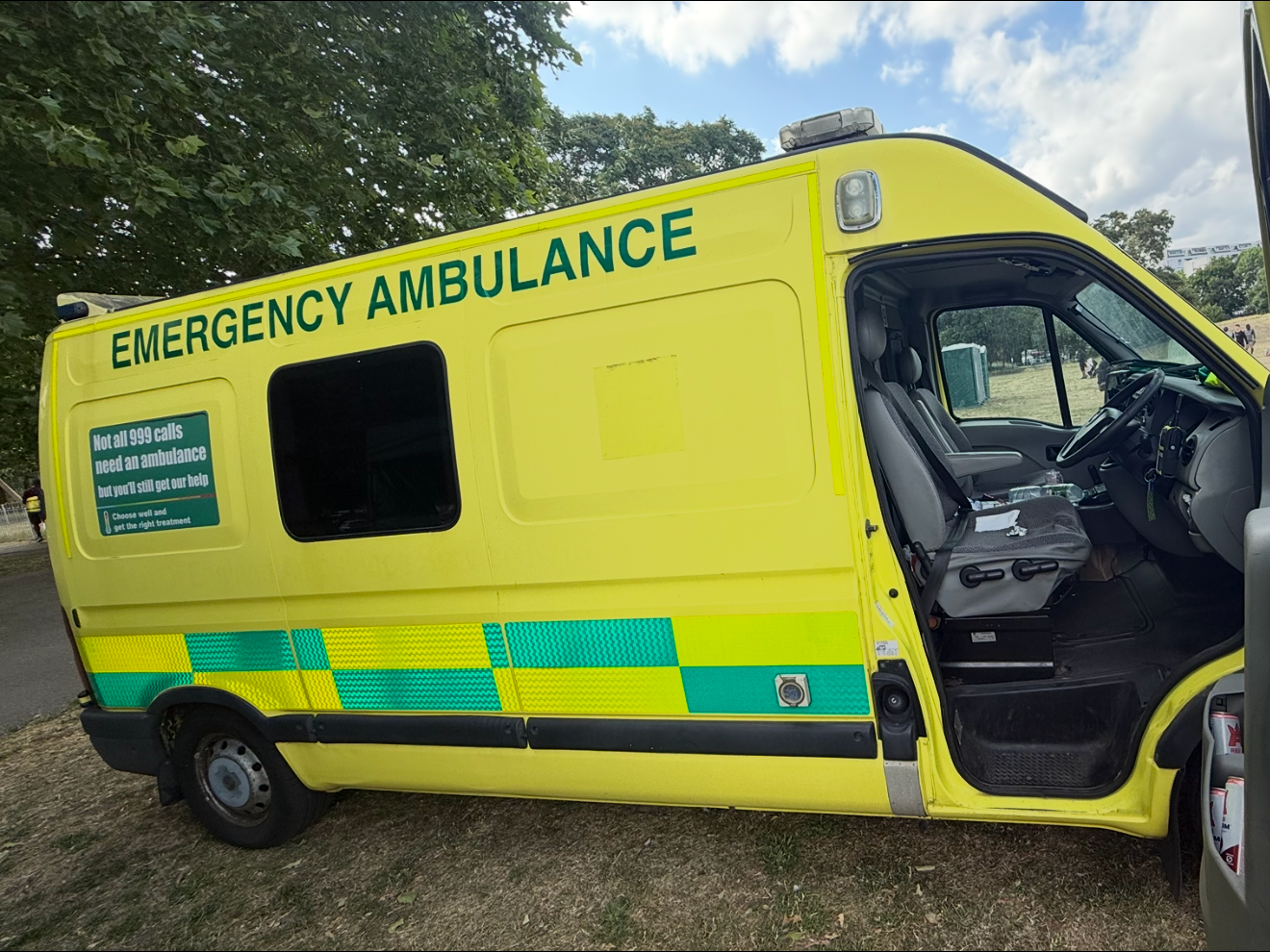 Yellow emergency ambulance vehicle with open driver's side door, parked outdoors near trees, with a blue sky and clouds in the background.