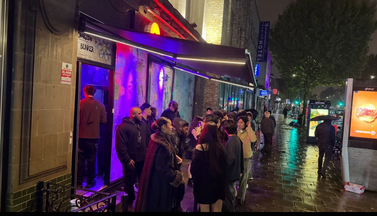 People waiting in line outside a venue on a rainy night, with some holding umbrellas, on a city sidewalk illuminated by colorful neon and street lights.