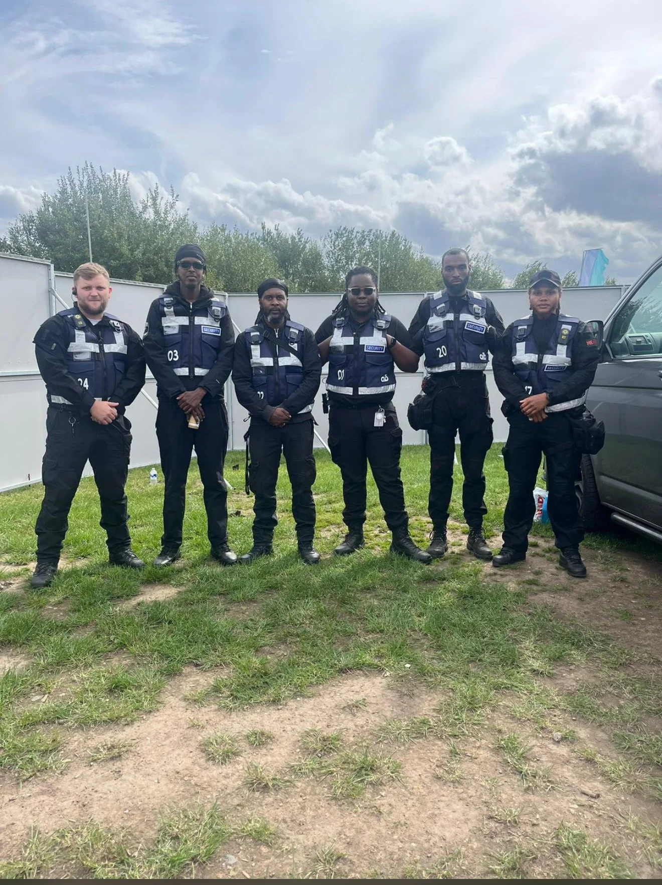Six security personnel standing outdoors on a grassy area, wearing black uniforms with reflective patches and gear, with cloudy sky and trees in the background.