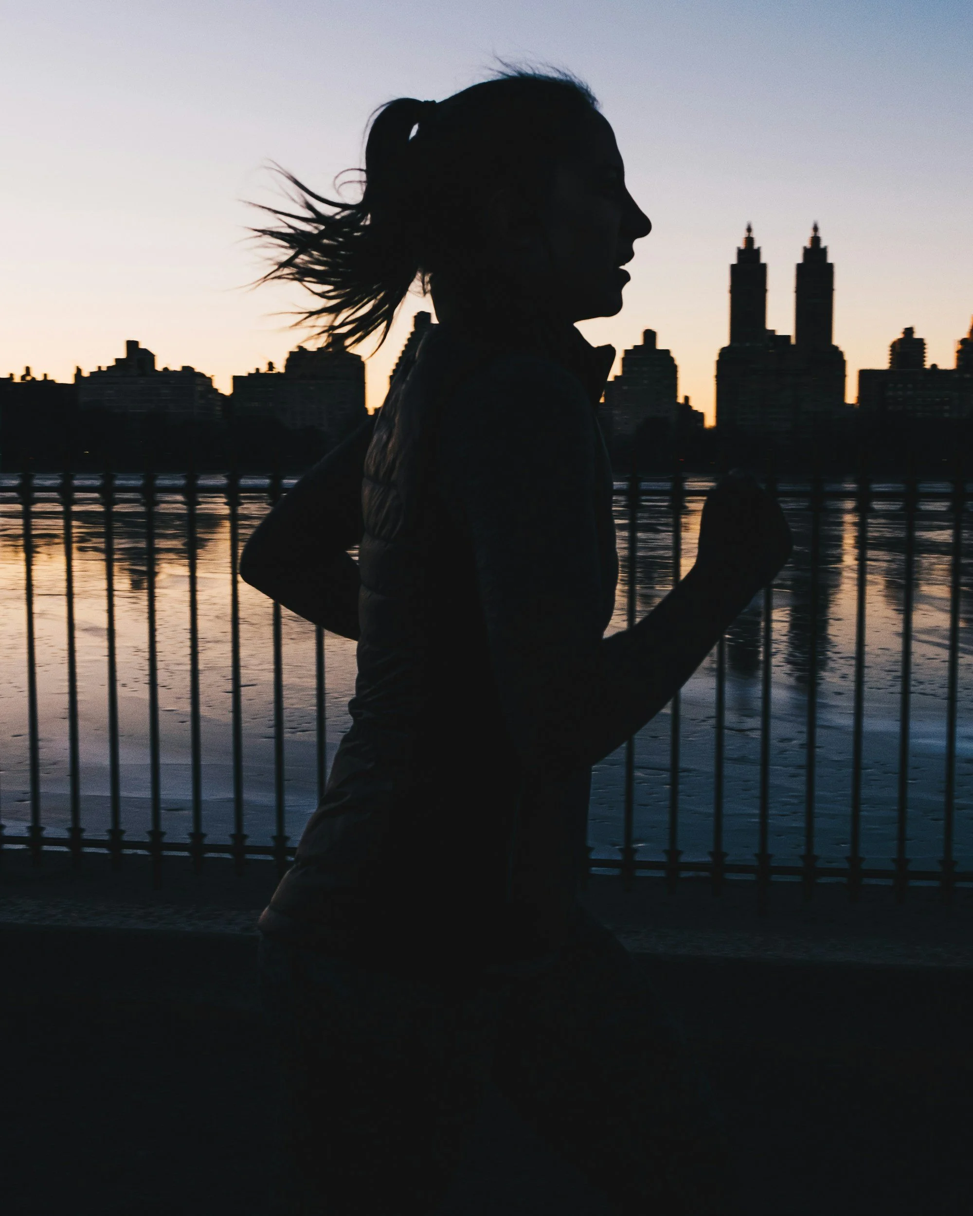 Silhouette of a woman running along a waterfront during sunset, with city skyline and tall buildings in the background.