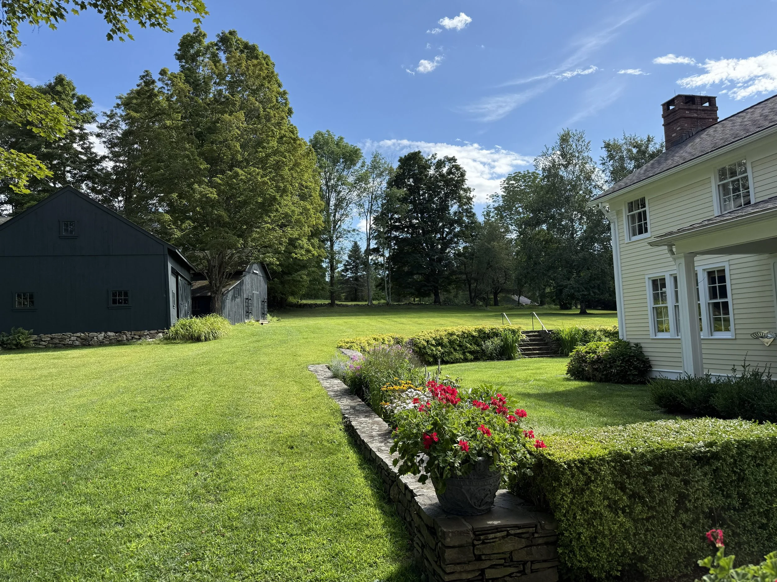 A sunny backyard with a white house, green lawn, colorful flowerbed, and black outbuilding surrounded by trees.