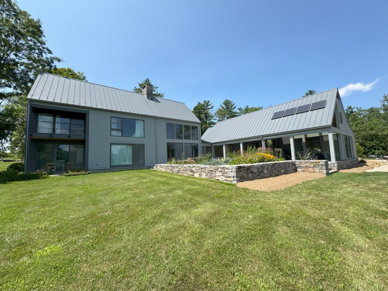 Modern house with gray exterior, metal roof, large windows, and a garden with a stone wall on a bright sunny day.