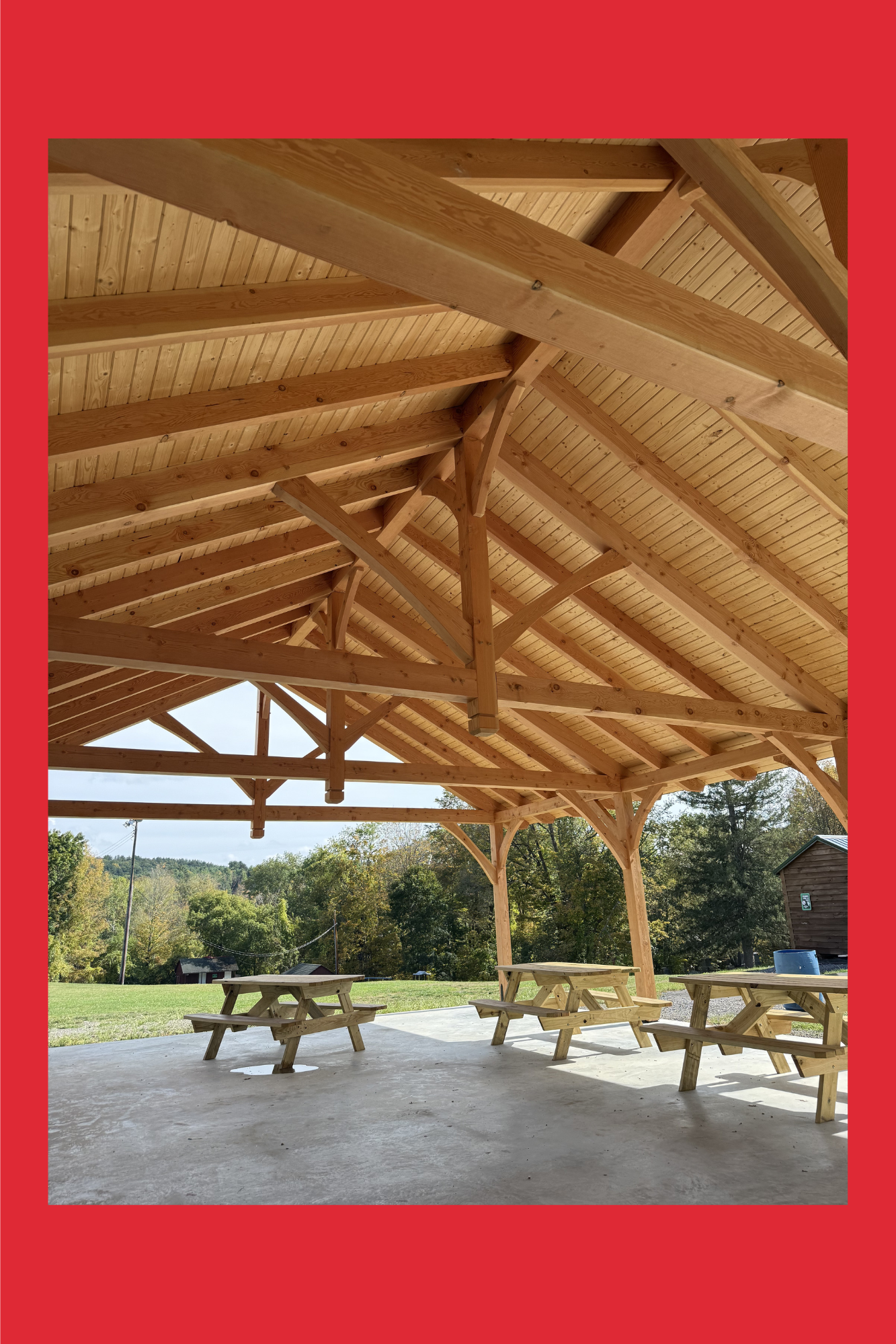 A wooden pavilion with a pitched roof and picnic tables underneath, set in a grassy outdoor area with trees in the background.