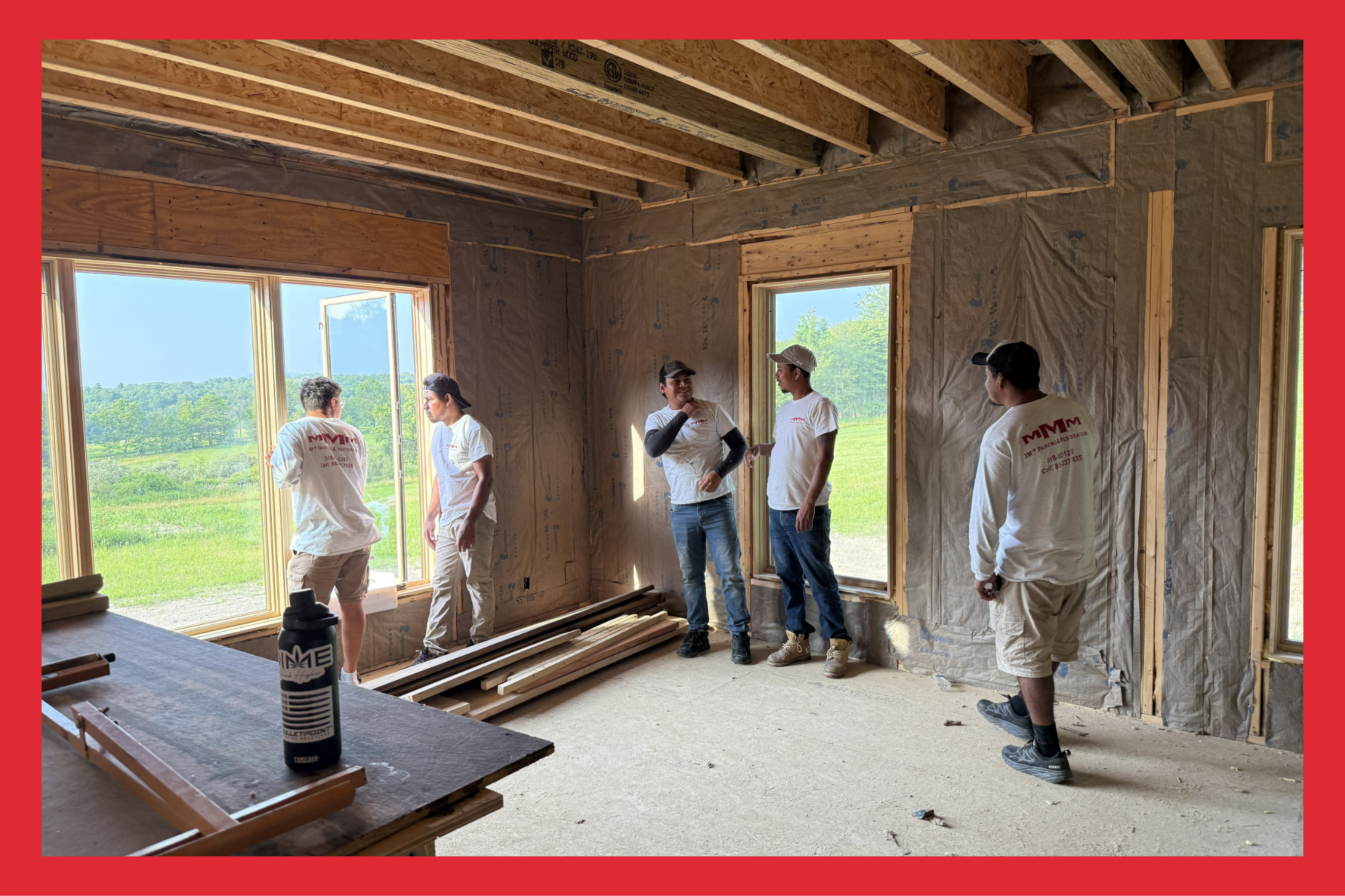 Group of construction workers inside a partially built house, with exposed wooden beams and insulation, discussing during daytime with green landscape visible outside through large windows.
