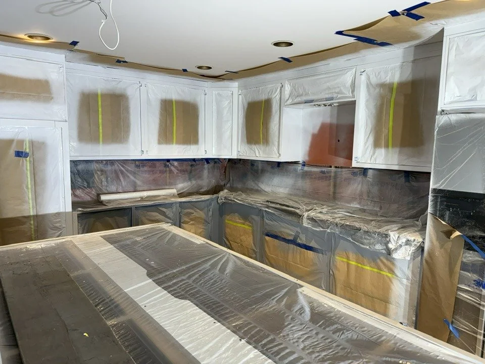 Kitchen under renovation with cabinets and countertops covered in protective plastic wrap and painter's tape.