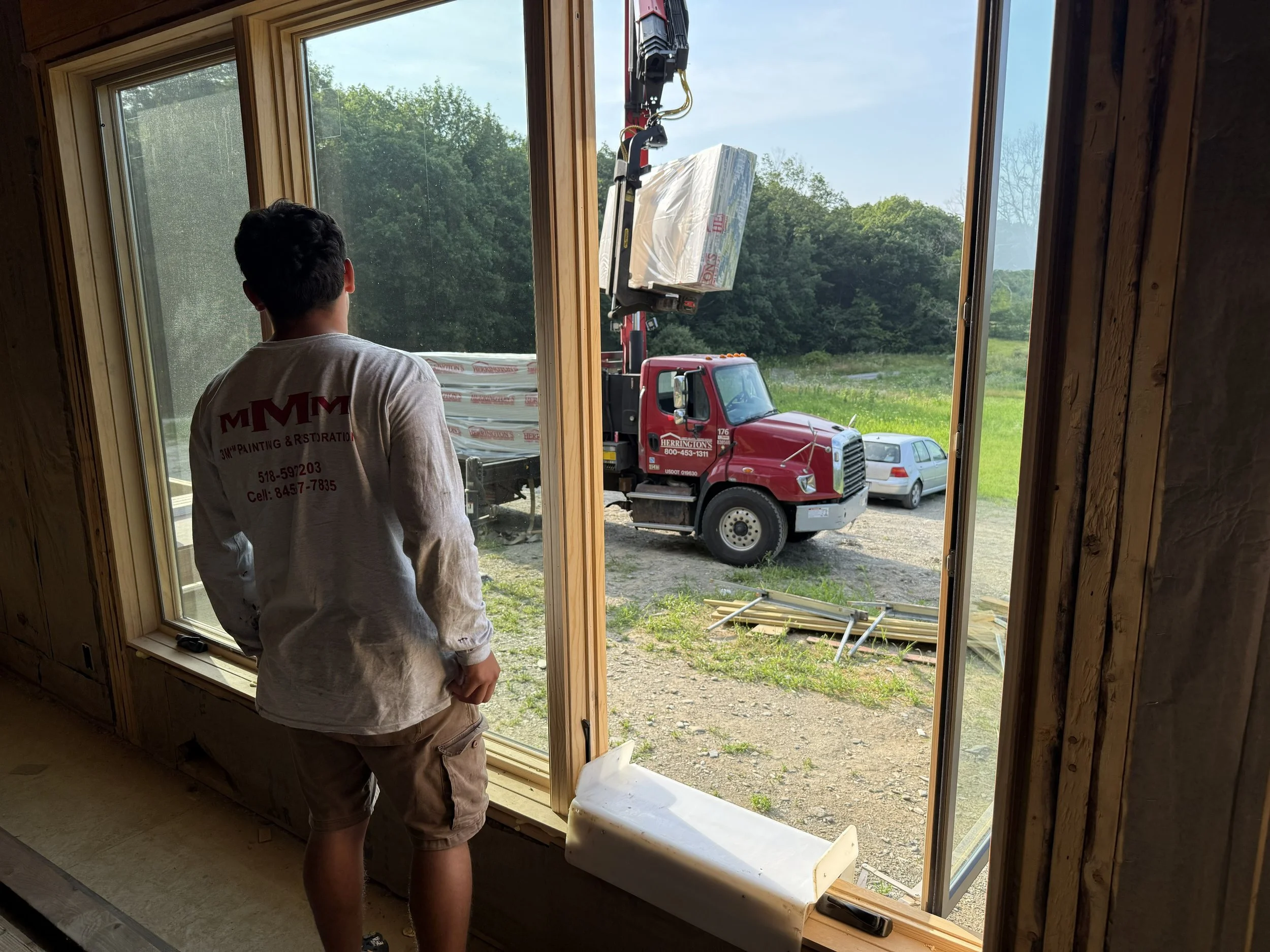 A man looking out of a large window at a construction site with a red crane truck lifting a package, and a white car parked outside on a grassy area.
