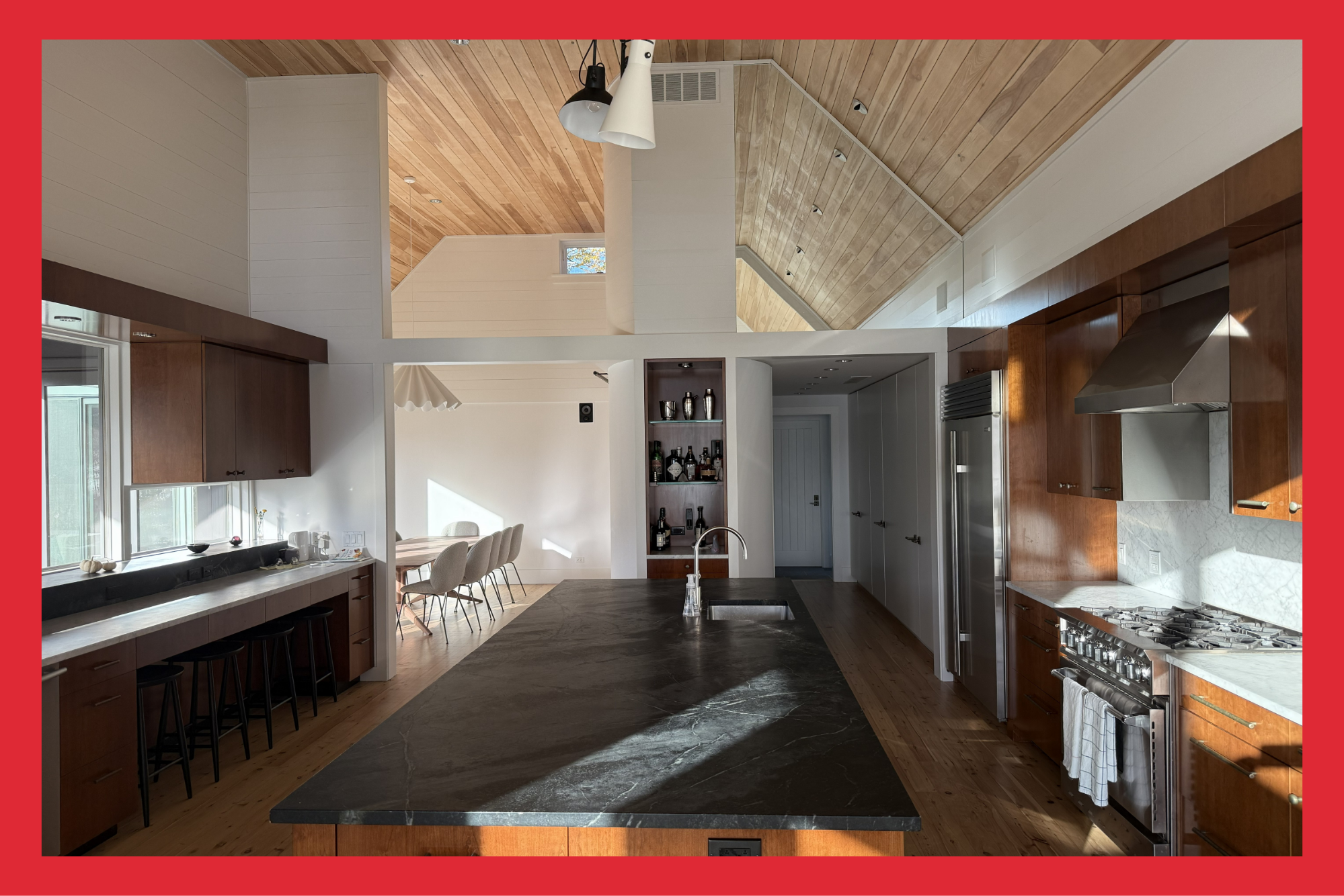 Modern kitchen with dark marble island, wooden cabinets, stainless steel appliances, and a adjacent dining area with beige chairs and a white wall